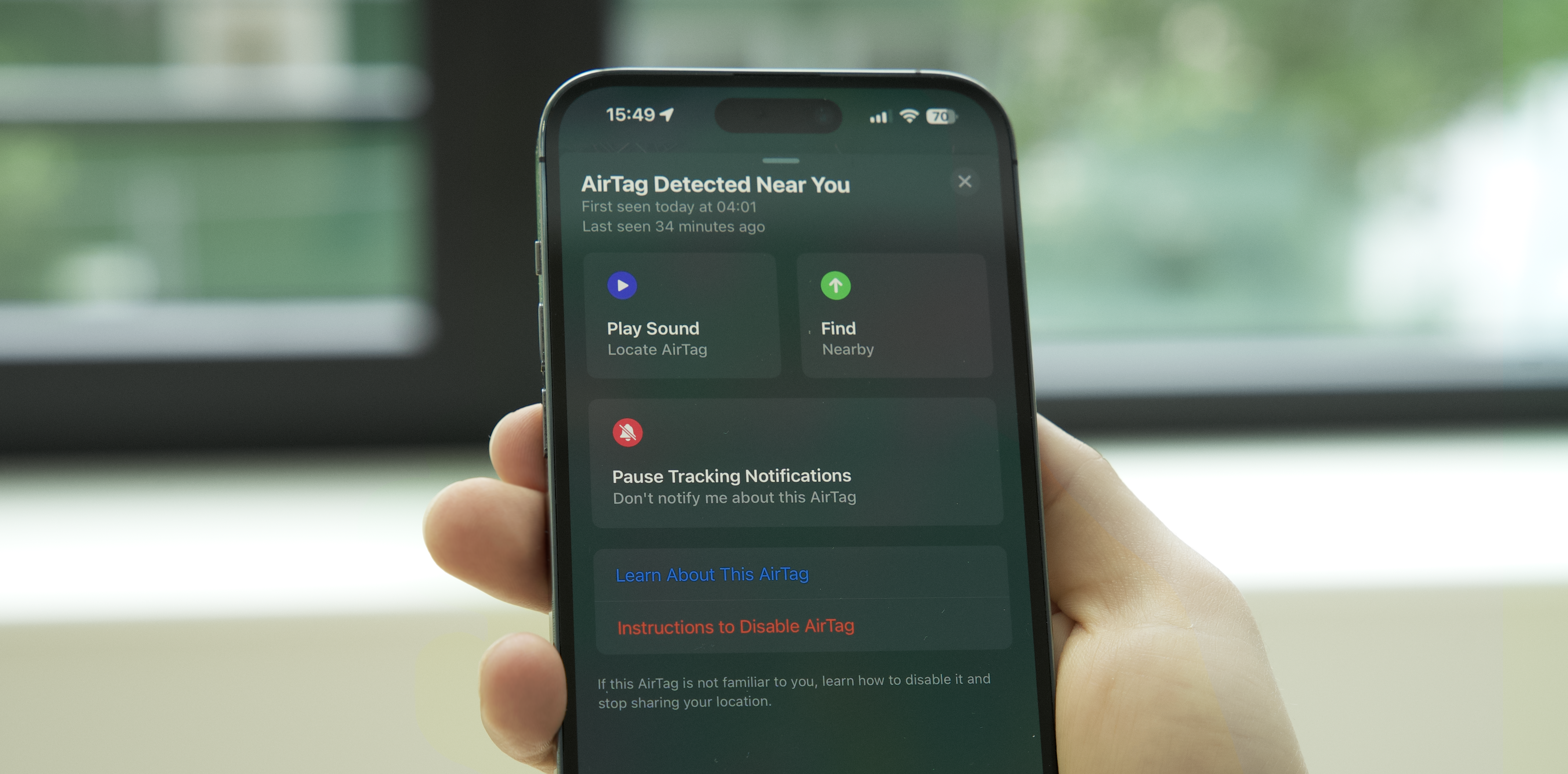 A hand holds an iPhone displaying a "AirTag Detected Near You" notification in the Find My app. The screen shows options to "Play Sound" and "Find" the AirTag, along with an option to "Pause Tracking Notifications" and links to "Learn About This AirTag" and "Instructions to Disable AirTag."